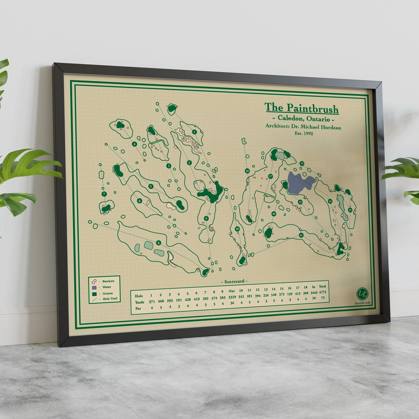 A framed vintage-style golf map print titled "The Paintbrush" leaning against a white wall on a light marble surface, with green plants partially visible at the sides.