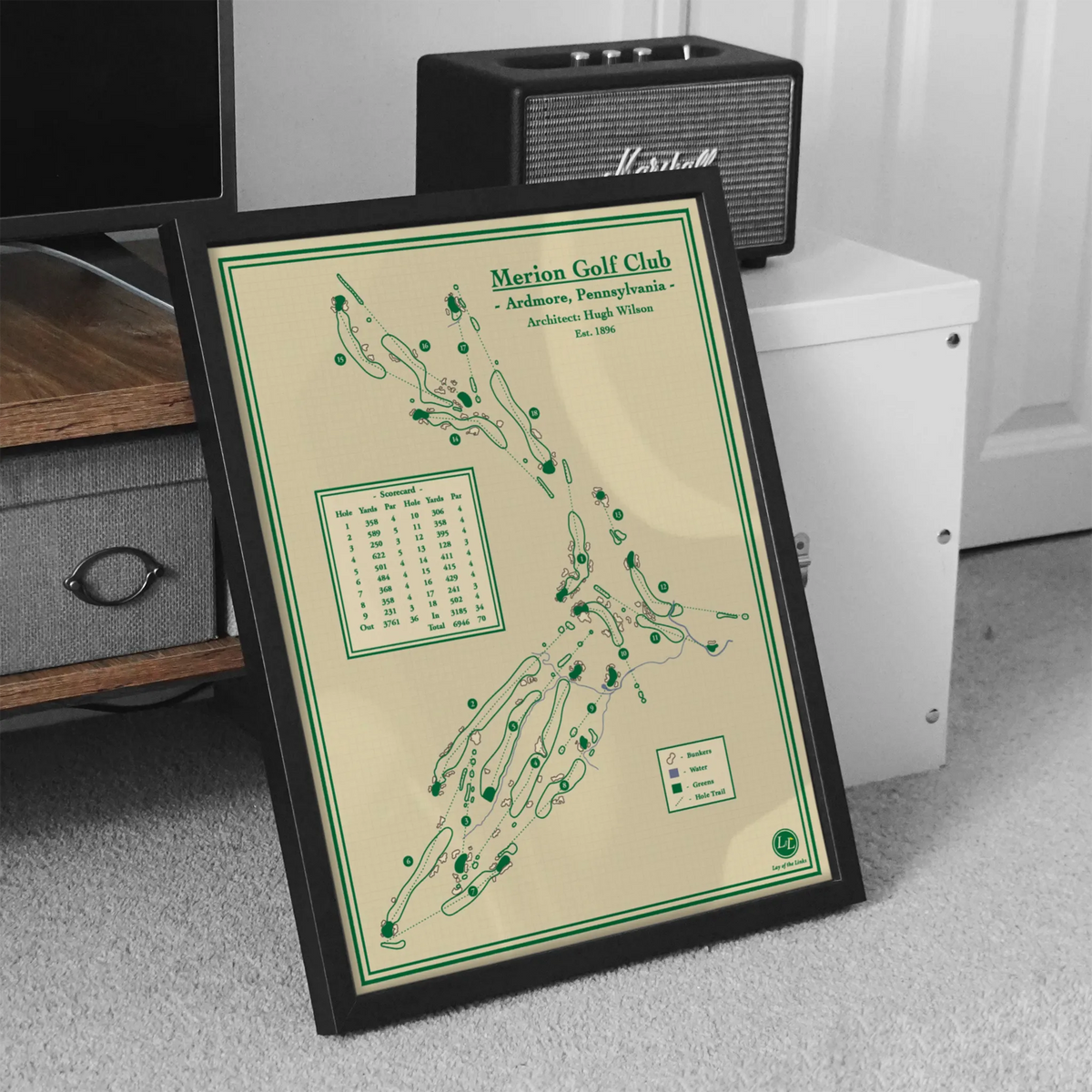 A framed vintage-style golf map print titled "Merion Golf Club" leaning against furniture in a room. The print has a cream background with green line art and text, enclosed in a black frame.