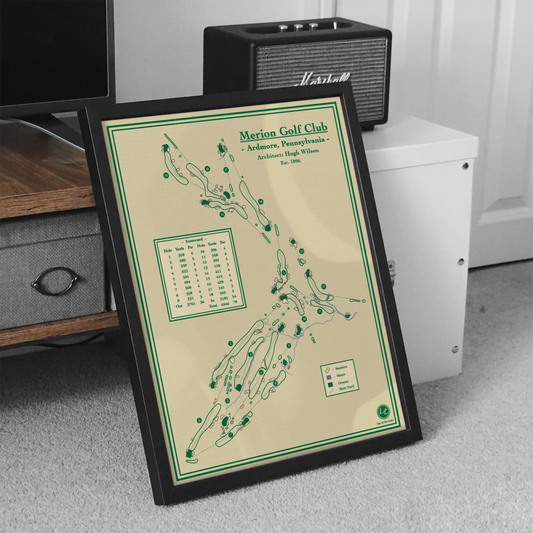 A framed vintage-style golf map print titled "Merion Golf Club" leaning against furniture in a room. The print has a cream background with green line art and text, enclosed in a black frame.