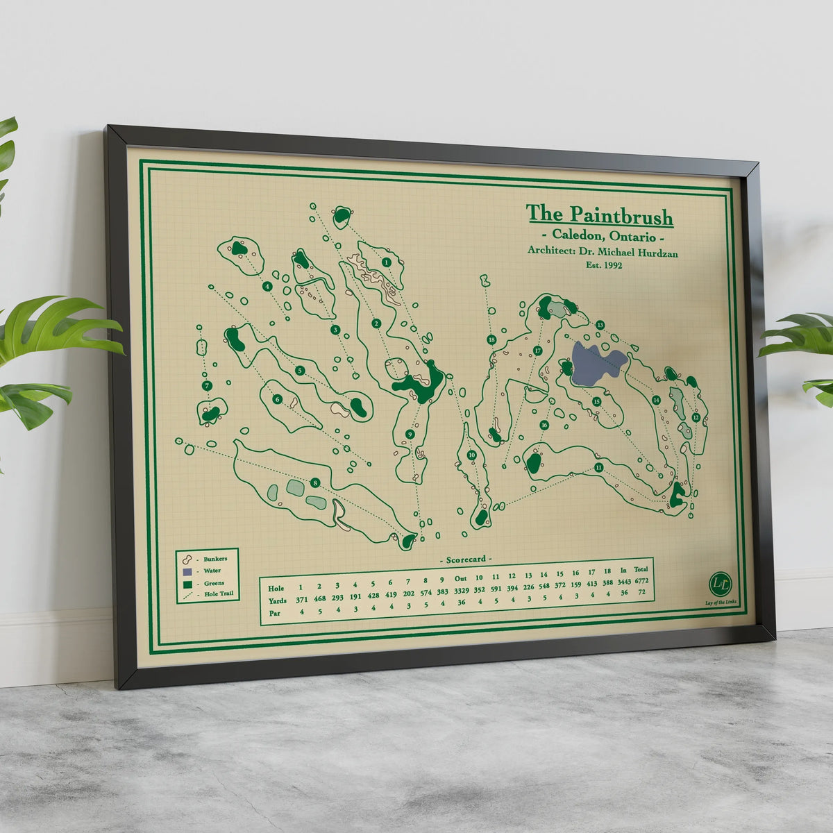 A framed vintage-style golf map print titled "The Paintbrush" leaning against a white wall on a light marble surface, with green plants partially visible at the sides.