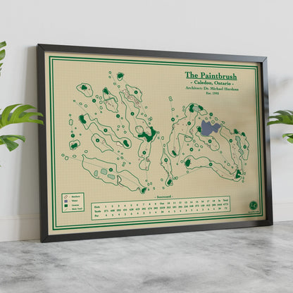 A framed vintage-style golf map print titled "The Paintbrush" leaning against a white wall on a light marble surface, with green plants partially visible at the sides.